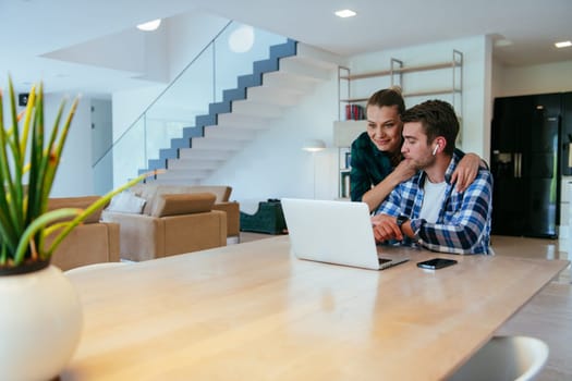 A young married couple is talking to parents, family and friends on a video call via a laptop while sitting in the living room of their modern house