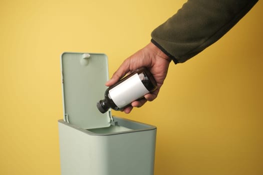 A hand placing a bottle into a green trash bin against a bright backdrop.