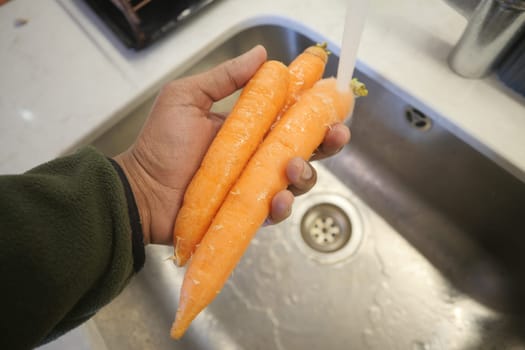 Hand holding clean, orange carrots over a stainless steel sink in kitchen.