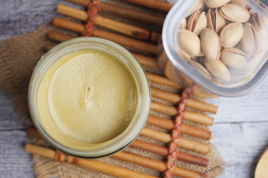 Pistachio spread sits in a clear glass jar beside shelled nuts on a mat.