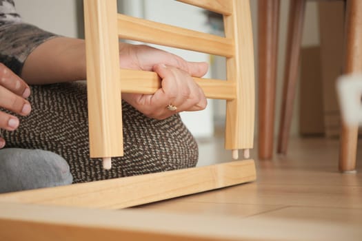 A person focuses on assembling a wooden chair on a warm wooden floor.