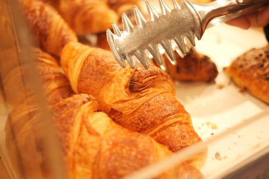 A hand uses tongs to select a golden croissant from a display.