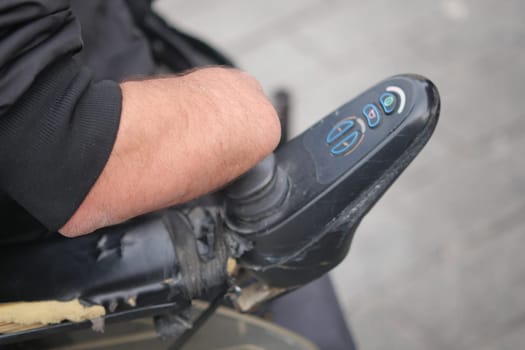 A hand controls a mobility scooter on a city sidewalk during the day.