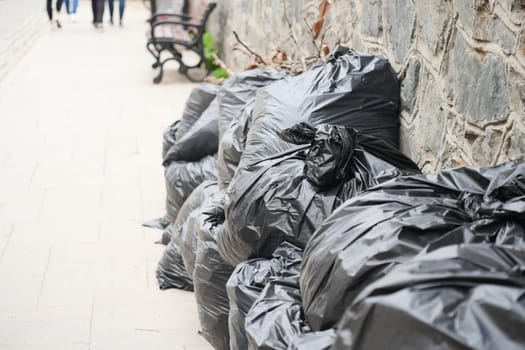 Uncollected trash bags line the sidewalk near a stone wall on a busy day.