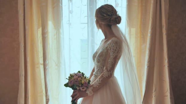 Beautiful blonde bride holds flower bouquet near the window.