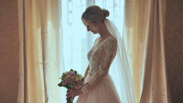 Beautiful blonde bride holds flower bouquet near the window.