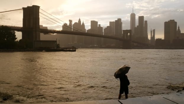 New York City, United States - 11 Sept 2023: Brooklyn Bridge rainy foggy day. Manhattan downtown skyline cityscape, World Trade Center. Man with umbrella, Dumbo Pebble Beach. Autumn river, fall in USA