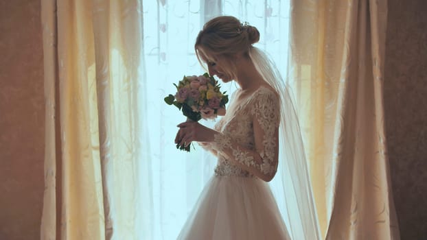 Beautiful blonde bride holds flower bouquet near the window.