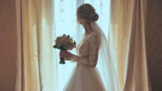 Beautiful blonde bride holds flower bouquet near the window.