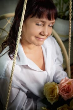 A middle-aged woman sits gracefully on a swing indoors, holding a bouquet of roses while smiling warmly in her stylish apartment.