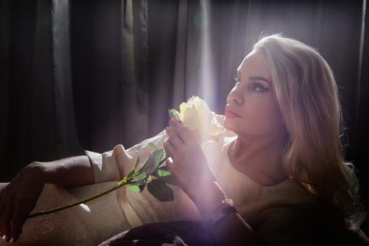 A middle-aged blonde woman poses elegantly with a rose in hand, capturing a serene moment in her home.