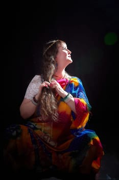 Woman with long hair dressed in a colorful sari enjoys a serene moment posing for an Indian style photoshoot in a studio setting.