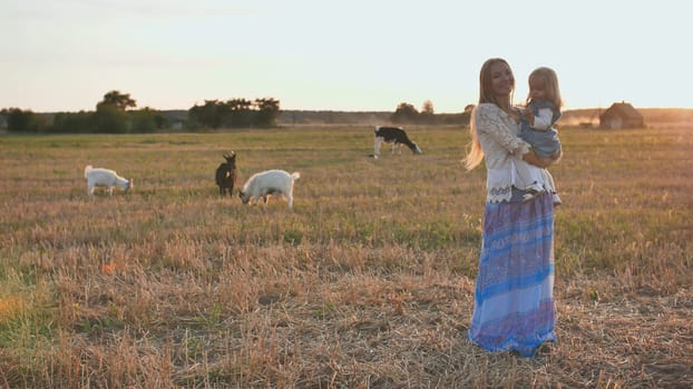 Mom holds a one-year-old daughter in her arms on a background of a village field with goats and a cow