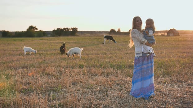 Mom holds a one-year-old daughter in her arms on a background of a village field with goats and a cow