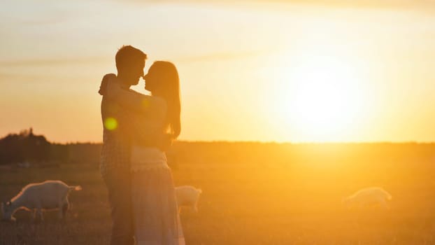 A silhouette of a girl approaching her beloved guy and hugs him against the backdrop of sunset and goats