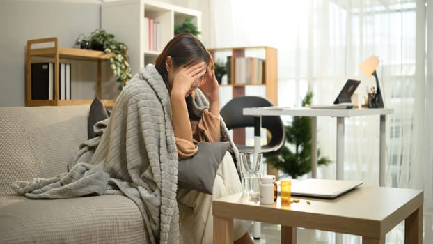 Young woman wrapped in blanket sitting on couch, suffering from headache or fever.