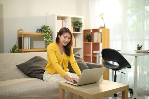 Cheerful asian woman holding a coffee mug and browsing on her laptop in living room.