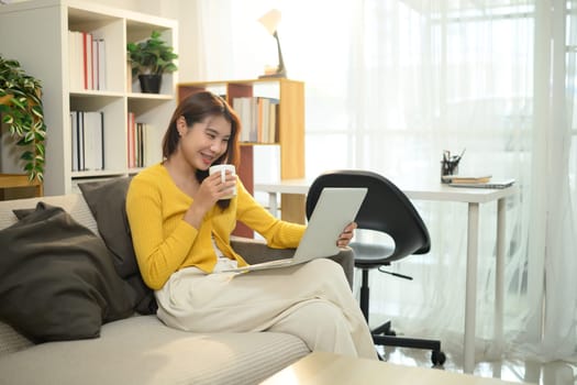 Cheerful asian woman holding a coffee mug and browsing on her laptop in living room.