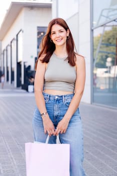 young adult woman in casual clothes standing in a sidewalk near of a mall carrying shopping bags and smiling happy looking at camera, concept of youth and urban lifestyle