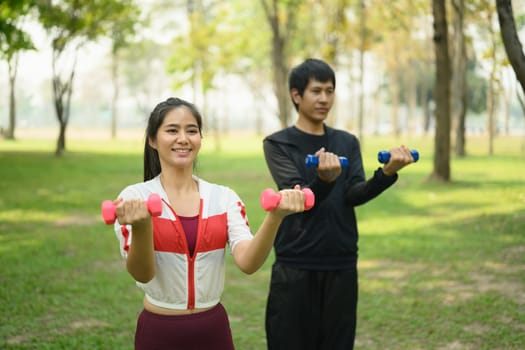 Active man and woman exercising with dumbbells together in a green park.