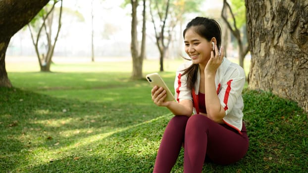 Smiling young woman sitting under a tree in a park, using her phone and wireless earbuds.