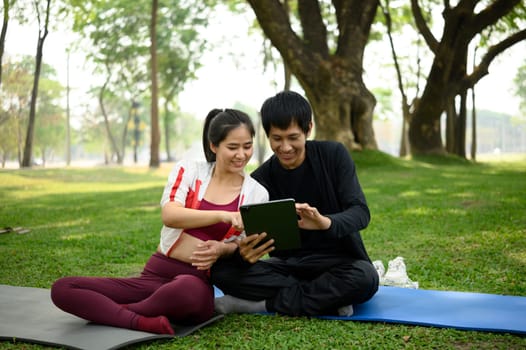 Asian couple checking workout plan on digital tablet, sitting on yoga mats in a sunny park.