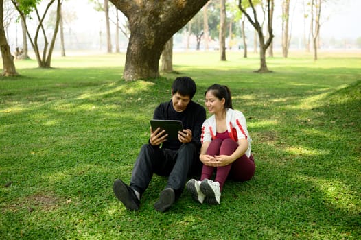 Asian couple checking workout plan on digital tablet, sitting on yoga mats in a sunny park.