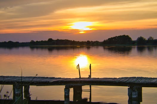 Sunset over a lake, a wooden footbridge leads into the water. Soft colors and reflections create a calm, idyllic scene. Ideal for travel dreams!