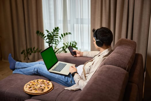 Young man sitting on sofa and eating pizza while working on laptop