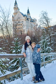 Enchanting winter lifestyle portrait capturing tender moment between mother and young boy exploring magical snow covered castle grounds. Elegantly dressed woman in contemporary winter outerwear holds hands with adorable child while standing on rustic wooden bridge surrounded by pristine winter scenery. Majestic historic castle with ornate towers provides stunning architectural backdrop against crystal clear blue sky. Natural authentic moment showcases family connection, winter tourism, fairytale atmosphere and European cultural heritage perfect for travel, tourism, parenting, and winter lifestyle marketing. Mother and Child Exploring Snowy Winter Castle Wonderland on Wooden Bridge Against Blue Sky with Historic Architecture and Snow Covered Landscape