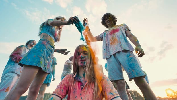 Young people having fun celebrating holi festival, pouring vibrant powder on girl's head, and enjoying outdoor festivities at sunset