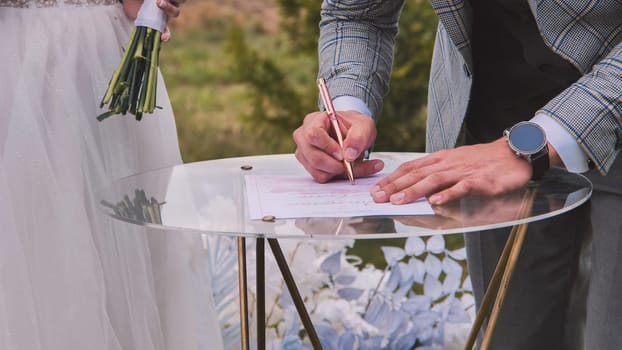 Groom signing marriage certificate on a glass table during an outdoor wedding ceremony, with the bride holding a bouquet