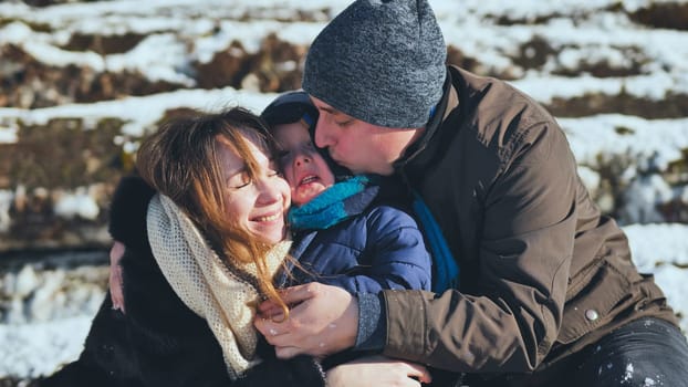 Parents hugging and kissing their joyful son on a chilly winter day, savoring the beauty of a picturesque snowy landscape together