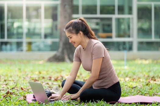 Embracing a modern lifestyle, a woman sits comfortably on her yoga mat, engaging with her laptop amid a serene outdoor backdrop.