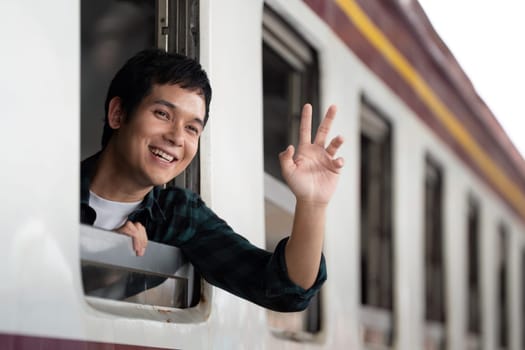 A young man happily waves from the window of a moving train, embracing the excitement of exploration and the thrill of adventure on his travels.