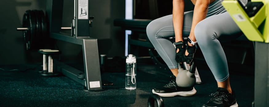 Asian woman holding and lifting a weight kettlebell in fitness gym, heavy kettle bell for strength training exercise in upper arm exercise to build muscle, Working out, healthy lifestyle concept
