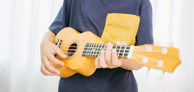 Close-up view of hands strumming a yellow ukulele, belonging to a young boy in a blue shirt. The white curtains in the background enhance the calm, creative vibe of the living room setting.