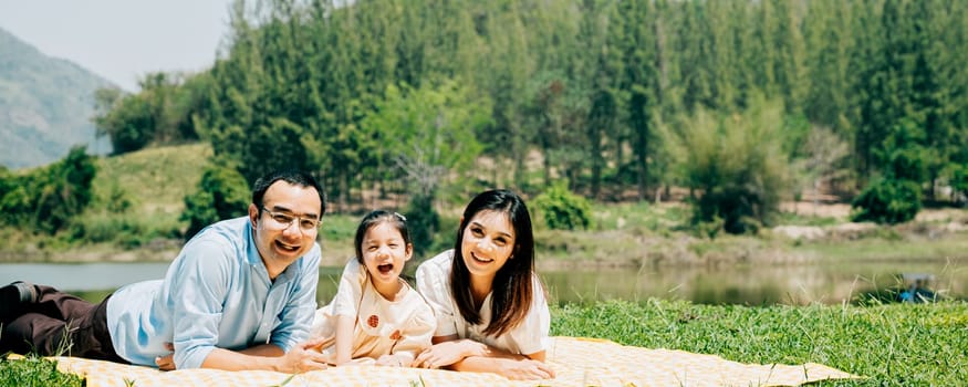 Asian family father mother and daughter lying on blanket spring meadow field grass in park cheerful sweet kid girl, parents and child picnic enjoying outdoors together, Family relaxation concept