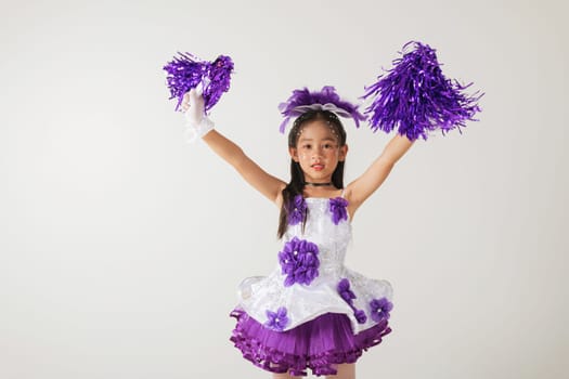 Cheerful young girl dressed as a cheerleader, holding purple pom-poms. Captured in a fun studio shot with a clean white backdrop. cheerleader kid