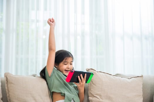 Happy Asian young girl using a tablet while sitting on a couch, excited child holding console playing video game raising hand look like win the games on sofa in living room at home online game concept