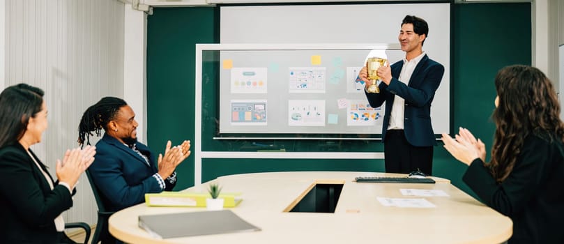 A successful businessman holds a performance trophy in a meeting room as the teamwork congratulates his achievement. This represents leadership, business success, and excellence in management.
