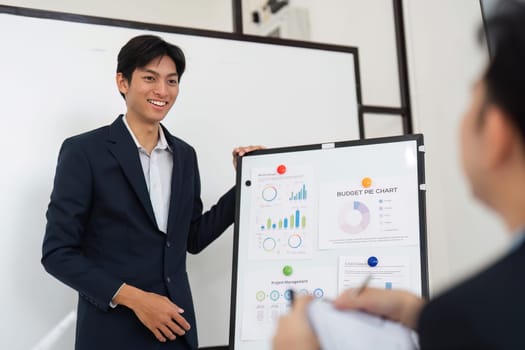 A confident male presenter explains a budget pie chart, engaging with team members who are attentively listening and taking notes. The setting emphasizes teamwork and effective communication.