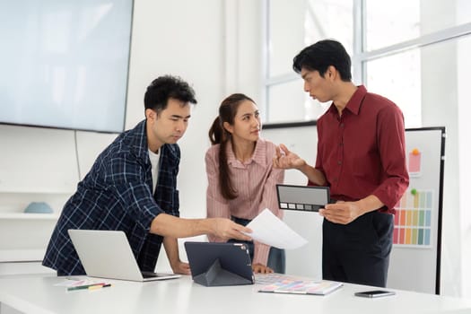 Three team members discuss innovative design ideas while analyzing color samples on their tablets and papers in a bright workspace, exemplifying teamwork and creativity.