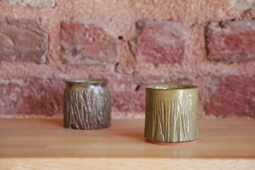 Two unique ceramic cups sit on a wooden table with a rustic brick wall behind.