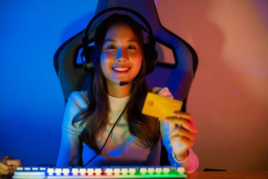 Asian gamer playing online video game and holding a credit card for payment game online, young woman in gaming headphones using computer for playing games at home, broadcast streaming live, Front view