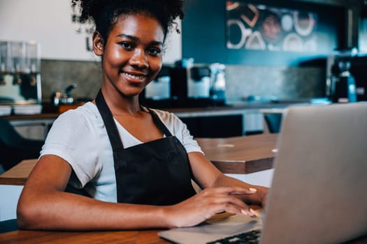 Portrait of a self-employed black woman barista at her coffee shop counter working on laptop. Confident entrepreneur managing finance bookkeeping in her own business.