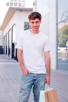 young adult man in casual clothes standing in a sidewalk near of a mall carrying shopping bags and smiling happy looking at camera, concept of youth and urban lifestyle