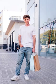 young adult man in casual clothes standing in a sidewalk near of a mall carrying shopping bags and smiling happy looking at camera, concept of youth and urban lifestyle, copyspace for text