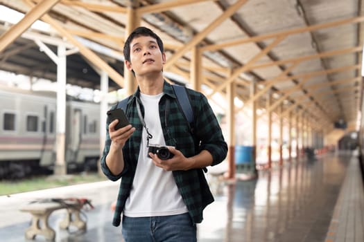 A young man stands in a train station, looking up with curiosity, phone in hand, ready to embark on his next adventure with camera slung over his shoulder.