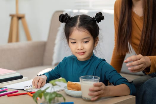 A young child sits at the table savoring a sandwich and milk while interacting with her mother, embracing the joyful moments of their morning routine before school.